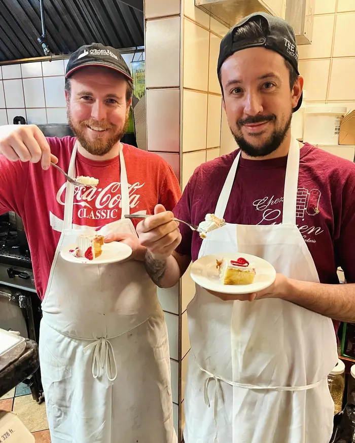 Two Chez Lucien cooks in aprons eating cake in the kitchen